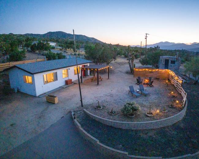A modern home with a fire pit and seating area is surrounded by desert landscaping, under a twilight sky.