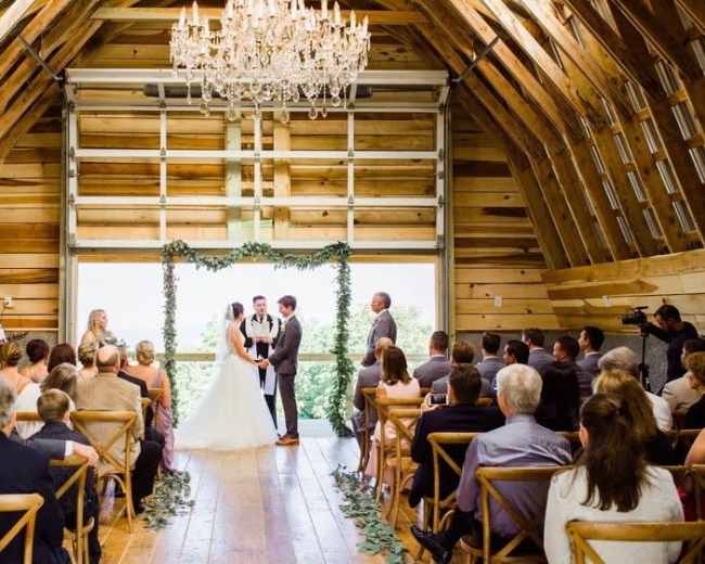 A couple stands at the altar in a rustic barn, surrounded by guests seated on wooden chairs, while a musician plays nearby.
