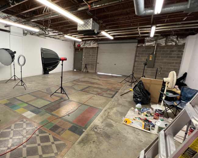 The image shows a photo studio setup featuring multiple lighting equipment, stands, and a partially cluttered floor in a spacious room with exposed ducts and a roll-up garage door.