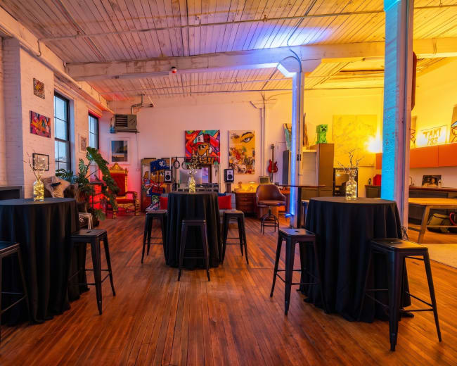 The image shows a well-lit, eclectic space with several tall tables covered in black cloth, surrounded by colorful artwork and furniture pieces.