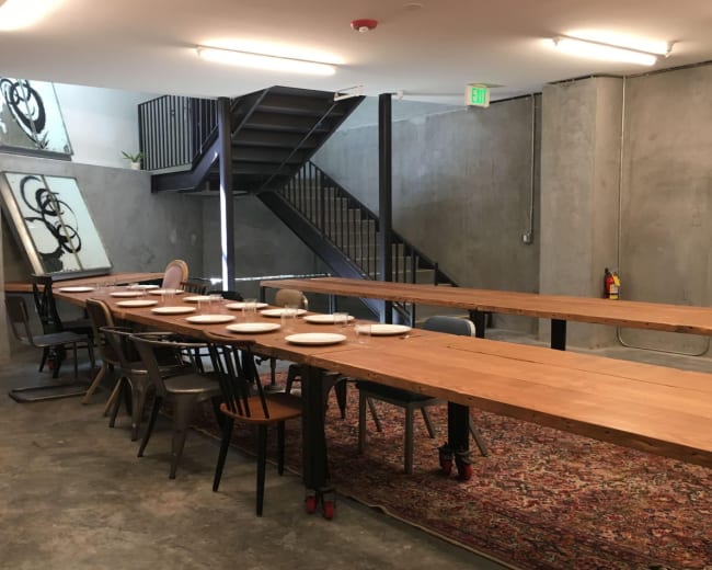 The image shows a large, elongated wooden table set with plates in a minimalistic room featuring concrete walls and a staircase in the background.