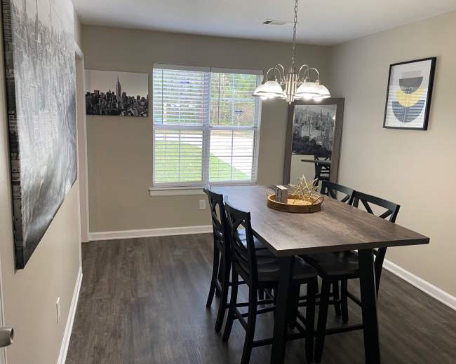 A dining area features a wooden table surrounded by black chairs, with light fixtures above and large windows letting in natural light.