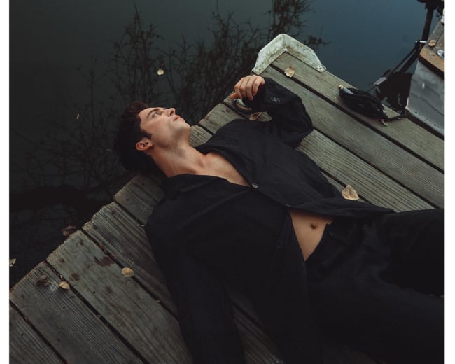 A man lies on a wooden dock near calm water, gazing upward with leaves scattered around him.