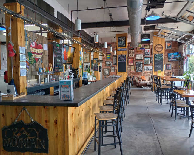 The image shows a rustic bar area with wooden furniture and decor, featuring a long bar counter and tables in a well-lit, open space.