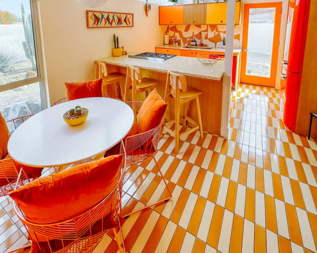 The image shows a brightly colored kitchen with orange cabinetry, a round white dining table surrounded by orange chairs, and a tiled floor in alternating stripes of orange and white.