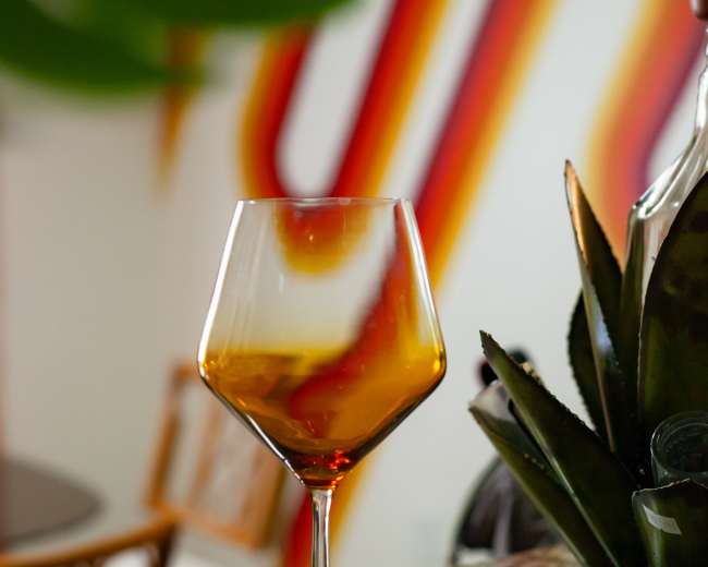 A half-filled glass of amber liquid stands on a speckled countertop in front of a colorful wall and a green plant.