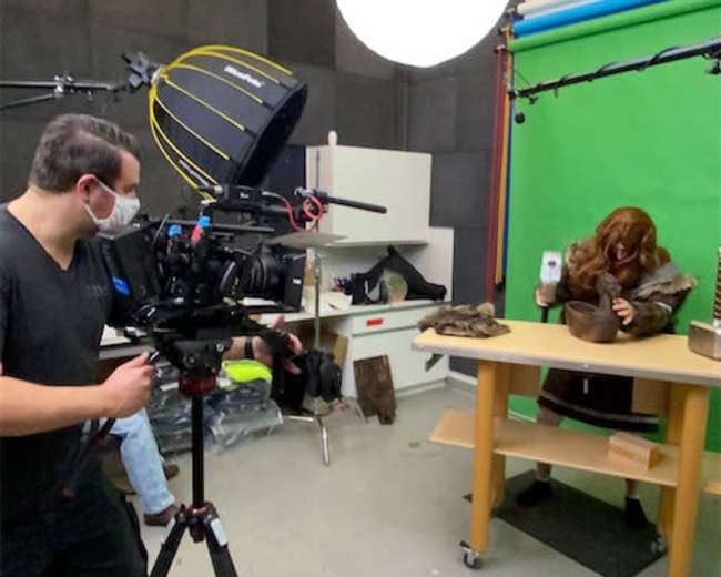 A person is filming with a camera in a studio, while a mannequin is seated at a table in front of a green backdrop.