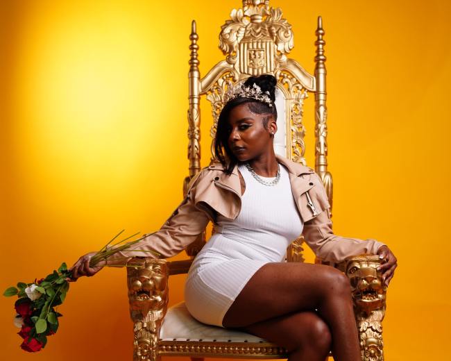 A person sits on an ornate golden throne against a vibrant yellow background, holding a bouquet of red roses.