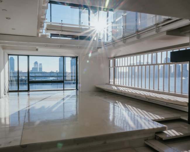 The image depicts a spacious, modern interior with large windows allowing natural light to illuminate the room and a view of a river and skyline beyond.