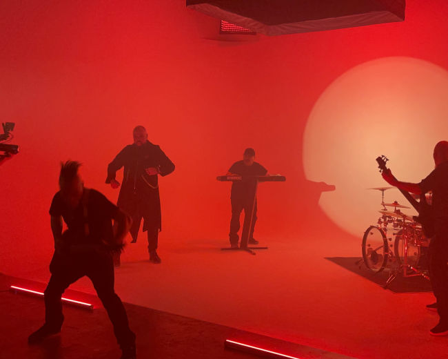 A music band performs in a dimly lit studio with red lighting and minimal stage equipment.