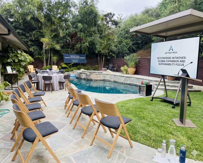 A set of chairs is arranged in front of a large screen displaying a presentation by a company, surrounded by a swimming pool and lush greenery in an outdoor setting.