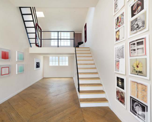 The image shows a modern interior with a staircase on the right, framed artwork on the left wall, and a large window allowing natural light to enter.