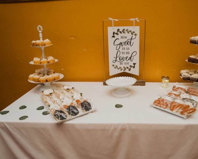 A dessert table features a sign that reads "How Sweet It Is to Be Loved by You," surrounded by various trays of cupcakes and decorated cookies.