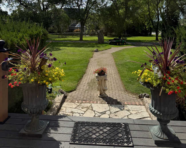 A pathway lined with flower pots leads through a green lawn surrounded by trees under a partly cloudy sky.