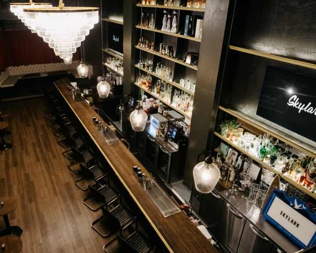 The image shows a modern bar with a long wooden counter, pendant lights, shelves stocked with various bottles, and a DJ setup in the background.