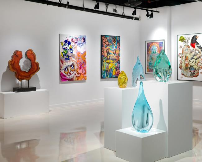 The image shows an art gallery with colorful paintings on the walls and several glass sculptures displayed on white pedestals.