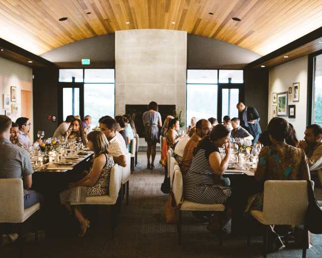 A group of diners sits at elegantly set tables in a modern restaurant with a wooden ceiling and large windows.