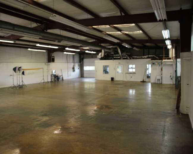 An empty industrial space with concrete flooring, large windows, and equipment for photography set up in one corner.