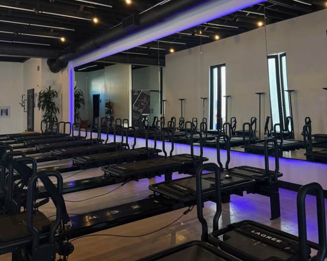 The image shows a modern fitness studio with multiple reformer machines arranged in rows, under a ceiling with exposed beams and bright overhead lighting.
