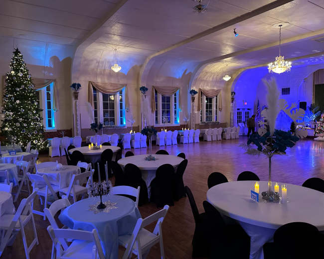 A decorated reception hall with circular tables covered in white and black linens, a large Christmas tree in the corner, and soft blue lighting illuminating the space.