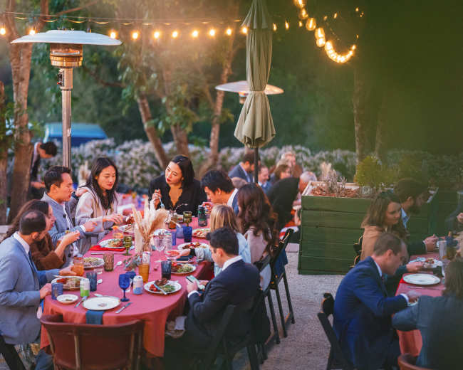 A group of people gather around outdoor tables enjoying a meal illuminated by string lights in a garden setting.