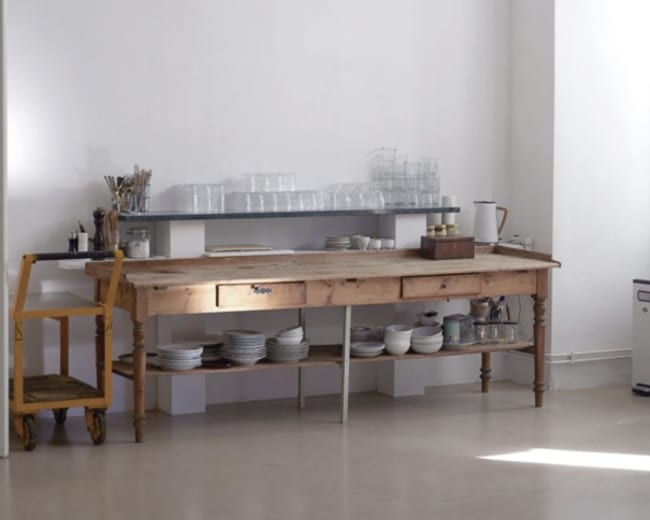 A wooden table with a drawer and a shelf above it holds glassware and plates in a bright, minimalist room.