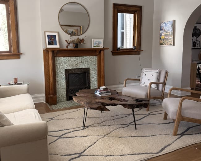 The image shows a living room featuring a fireplace with a circular mirror above it, a unique wooden coffee table, two armchairs, and a light-colored sofa, all arranged on a patterned rug.