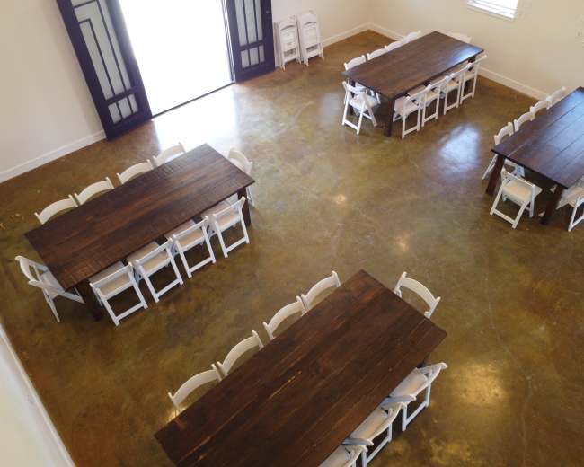 The image shows a well-lit room with four wooden tables arranged in a central layout, surrounded by white folding chairs.
