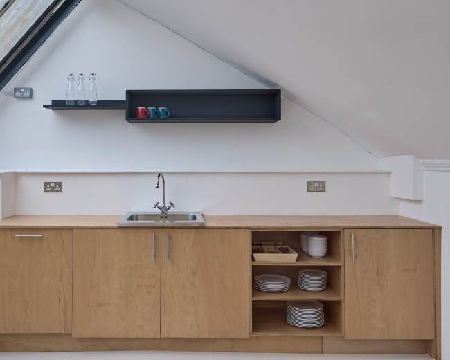 The image shows a modern kitchen area with wooden cabinetry, a stainless steel sink, and a shelf displaying bottles and colorful mugs.
