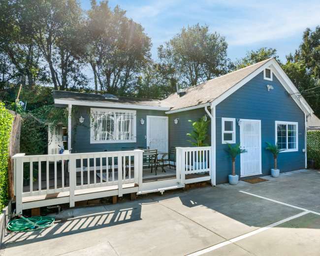 A blue cottage with a front porch and a small seating area is surrounded by greenery and a concrete driveway.