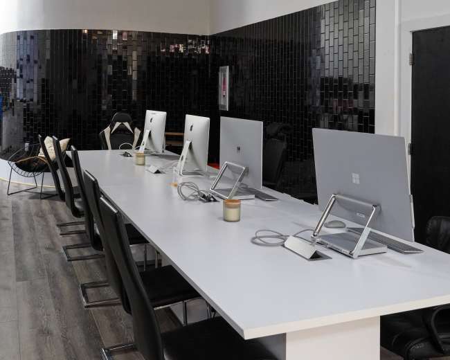 A row of computers is set up on a white table in a modern office space, accompanied by black chairs and a dark wall in the background.