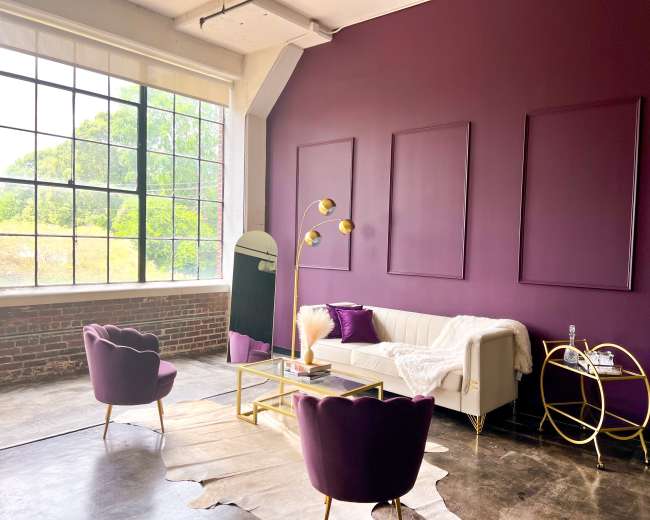 A modern living room features a white sofa, two purple chairs, a gold coffee table, and a large window with a view of greenery outside.