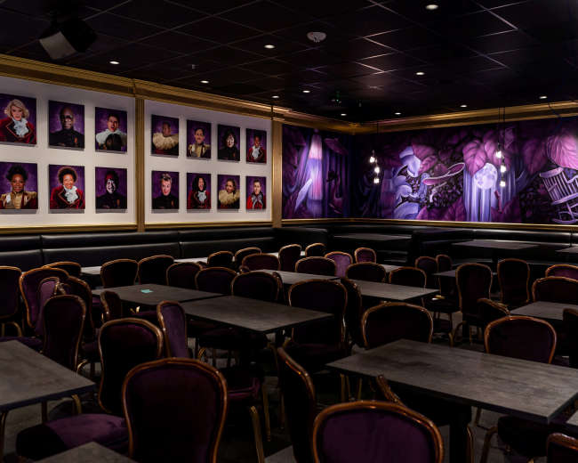 The image shows a dimly lit interior of a restaurant or café featuring purple decor, several tables, and a mural on the wall with portraits.