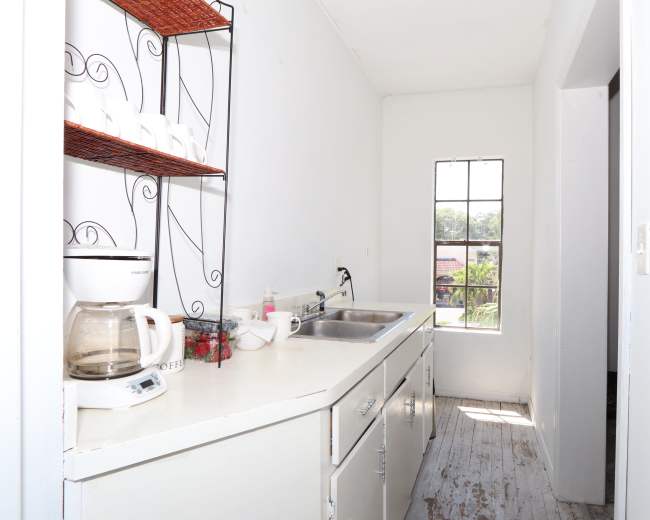 A narrow kitchen area with a sink, coffee maker, and open shelving holding cups, illuminated by natural light from a window.