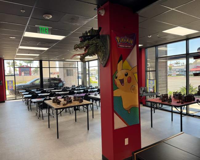 The image shows an indoor space decorated with Pokémon memorabilia, featuring tables set up for an event and large windows allowing daylight to enter.