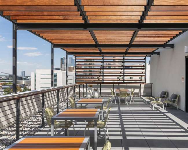 The image shows a modern rooftop terrace featuring wooden slatted awnings, tables with orange accents, and a view of city buildings in the background.