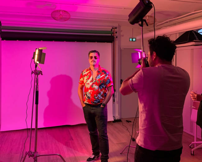 A man in a floral shirt poses for a photo while another person adjusts the camera under pink lighting.