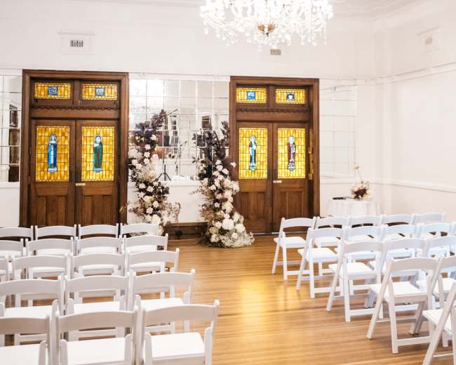 A spacious event room features rows of white chairs facing ornate stained glass doors adorned with floral arrangements.