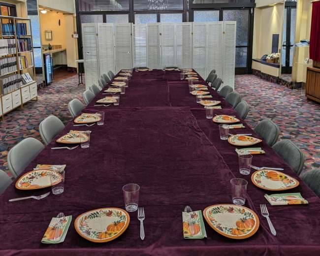 A long, rectangular dining table is set with plates and utensils, draped in a purple tablecloth, beneath a chandelier in a spacious room divided by folding screens.