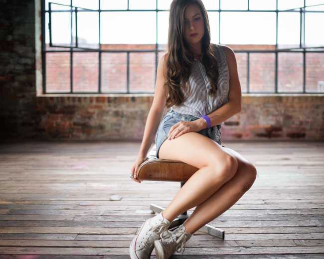 A woman sits on a wooden stool in a spacious, industrial-style room with large windows.