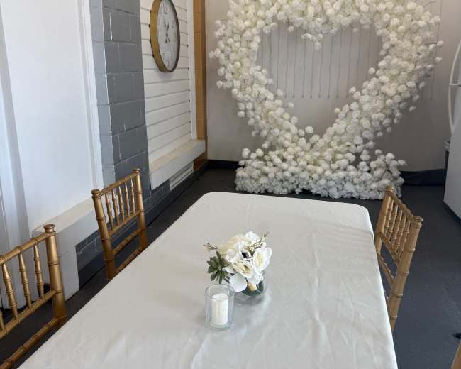 A table with a white tablecloth is set up in front of a heart-shaped floral arrangement, with a small floral centerpiece and a candle on the table.