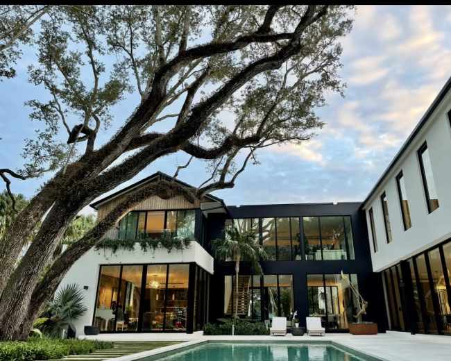 The image depicts a modern house with large glass windows, surrounded by greenery and a swimming pool in the foreground.