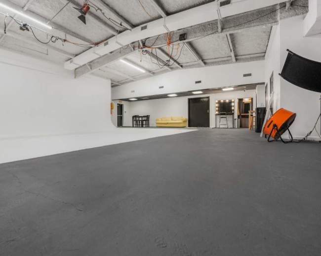 The image shows a spacious, empty studio with a white backdrop, gray flooring, and various furniture pieces, including a yellow couch and a mirrored vanity area.