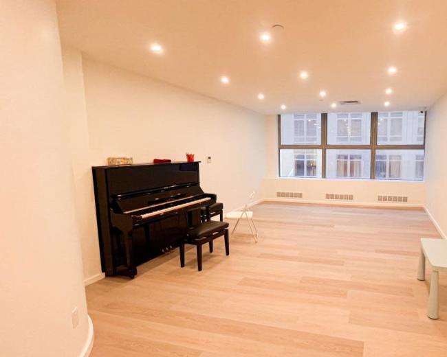 The image shows a spacious, empty room with a black piano, a chair, and a small light blue table, illuminated by recessed ceiling lights.