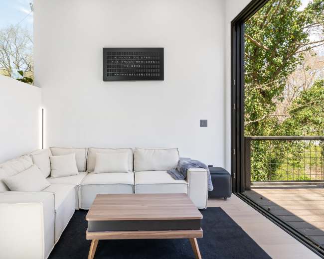 A modern living room features a white sectional sofa, a wooden coffee table, and large windows overlooking greenery.