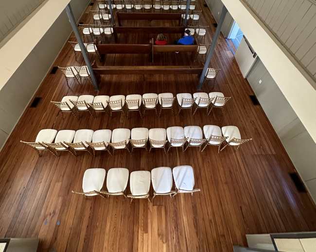 The image shows an empty, spacious indoor hall with rows of white chairs arranged on a wooden floor, flanked by balcony seating above.