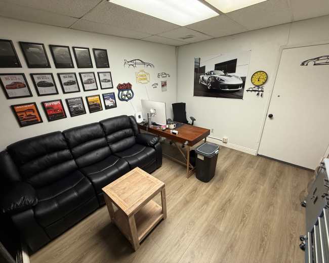 The image shows a small office or lounge area featuring a black leather sofa, a wooden table, and wall decorations that include framed awards and car-related artwork.
