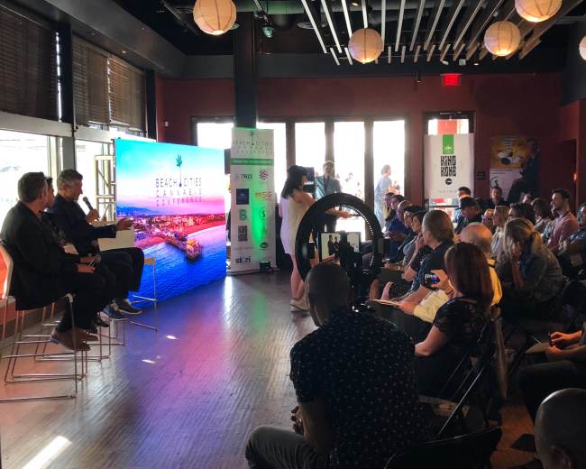 A panel discussion is taking place in a brightly lit venue, with an audience seated in front of a large screen displaying "Beach cities & cannabis."