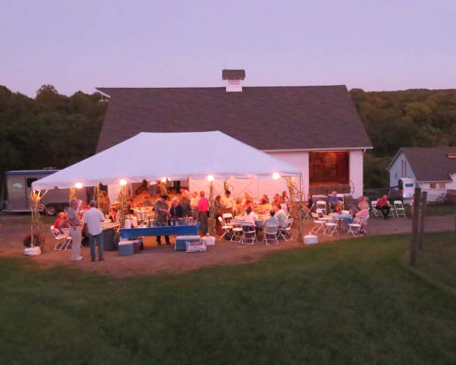 A large white tent with chairs and tables is set up outside a barn, with people gathered for an evening event as dusk falls.