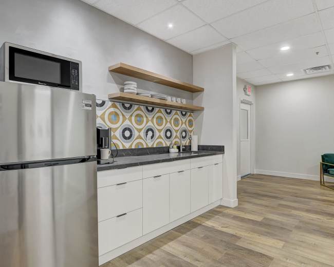 A modern kitchen area featuring a stainless steel refrigerator, microwave, and a patterned backsplash with decorative shelving above a counter.